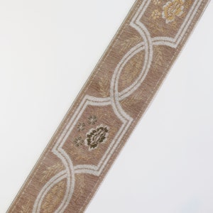 May include: A beige trim with a white and gold floral pattern. The trim has a repeating design of a stylized flower and a curved, interlaced pattern.
