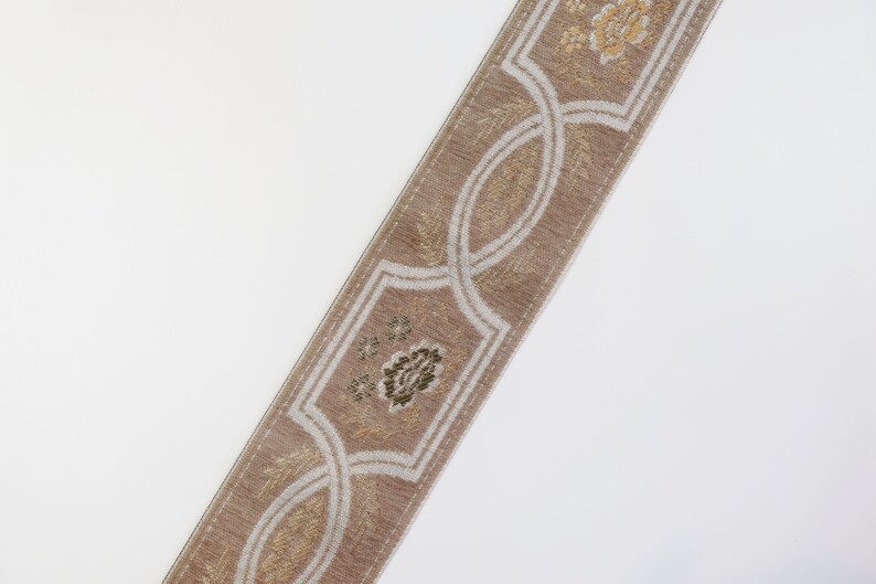 May include: A beige trim with a white and gold floral pattern. The trim has a repeating design of a stylized flower and a curved, interlaced pattern.