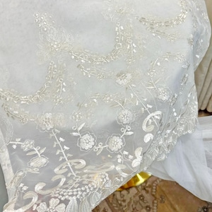 May include: Close-up of a sheer white fabric with intricate floral embroidery. The delicate design features detailed flowers, leaves, and vines, creating a textured, elegant look. The fabric's edge has a scalloped trim, adding to its refined aesthetic.