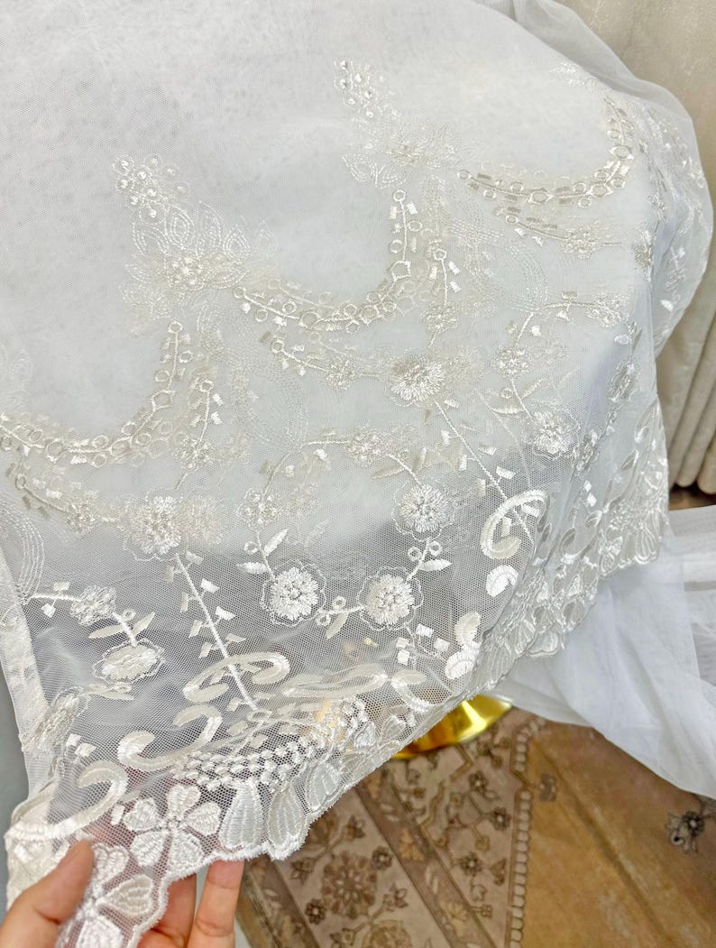 May include: Close-up of a sheer white fabric with intricate floral embroidery. The delicate design features detailed flowers, leaves, and vines, creating a textured, elegant look. The fabric's edge has a scalloped trim, adding to its refined aesthetic.