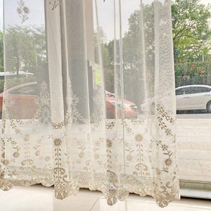 May include: Sheer, cream-colored curtain with intricate floral embroidery. The curtain features a delicate, lacy border and repeating floral patterns. The design is visible against the backdrop of a window with a view of trees and cars.