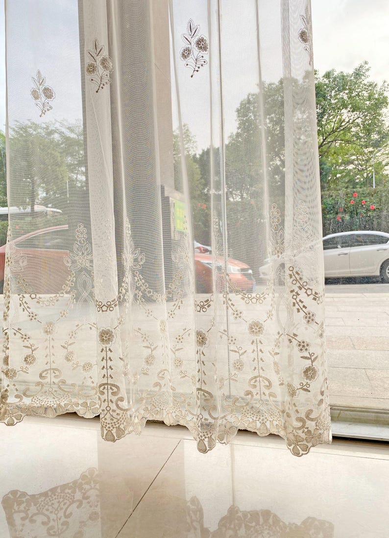 May include: Sheer, cream-colored curtain with intricate floral embroidery. The curtain features a delicate, lacy border and repeating floral patterns. The design is visible against the backdrop of a window with a view of trees and cars.