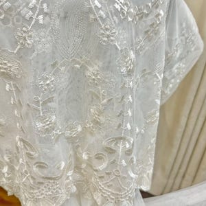 May include: Close-up of white lace fabric with intricate floral and geometric patterns. The delicate lace is draped, showcasing the detailed embroidery and texture. The fabric appears to be a light, airy material, suitable for bridal or decorative purposes.