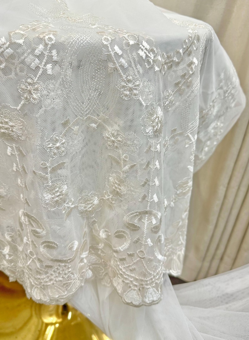 May include: Close-up of white lace fabric with intricate floral and geometric patterns. The delicate lace is draped, showcasing the detailed embroidery and texture. The fabric appears to be a light, airy material, suitable for bridal or decorative purposes.