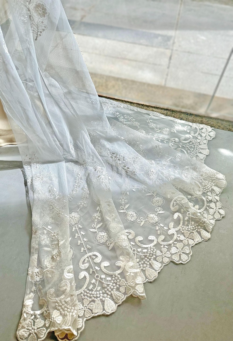 May include: White lace fabric with floral and leaf patterns. The delicate material is draped, showcasing the intricate embroidery along the edges. The fabric is likely used for decorative purposes, such as curtains or clothing.