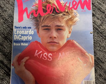 May include: A vintage magazine cover featuring Leonardo DiCaprio holding a red lip-shaped balloon that says "KISS ME." The magazine is titled "Interview" in large pink letters, with text that reads "June 1994."
