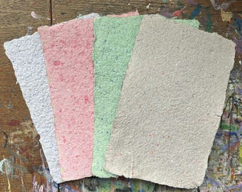 May include: A collection of handmade paper sheets in various colors. The sheets include white, pink, green, and beige, each with a textured surface and irregular edges. The paper appears to be made from recycled materials, with small flecks of color throughout.