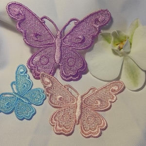 Puede incluir: Parches bordados con forma de mariposa en tonos morado, azul y rosa, sobre una superficie blanca. Tambi&eacute;n se muestra una flor de orqu&iacute;dea blanca con centro amarillo. Los parches de mariposa tienen dise&ntilde;os detallados en las alas.