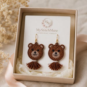 Könnte beinhalten: Ein Paar braune, bärenförmige Stickohrringe mit goldfarbenen Haken und braunen Garnquasten. Die Ohrringe sind auf einer weißen Karte mit der Aufschrift MyStitchMuse Embroidery Jewelry befestigt und befinden sich in einer hellen Geschenkbox.