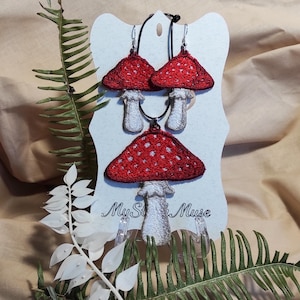 May include: A set of mushroom-themed jewelry, including earrings and a pendant. The mushroom caps are red with white spots, and the stems are white. The jewelry is displayed on a light blue card with the text "My St Muse".