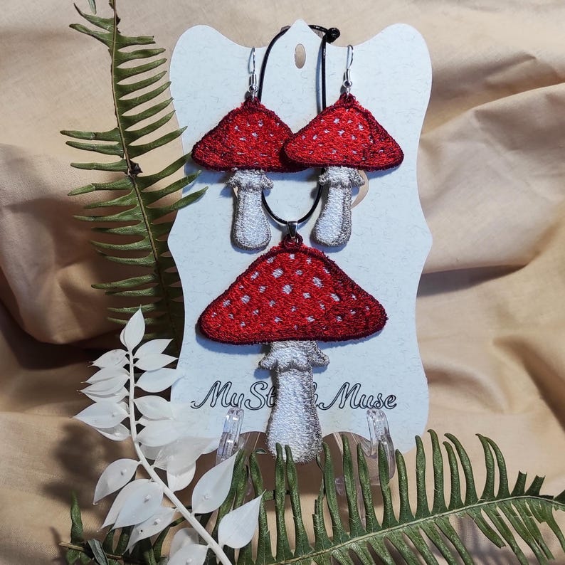 Set of FSL machine embroidery designs featuring red and white Amanita mushrooms used as earrings and pendant on a white display card with the MyStitchMuse logo, surrounded by decorative ferns.