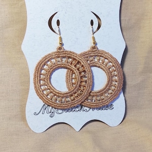 May include: A pair of gold-colored, crocheted hoop earrings with small beads. The earrings are displayed on a white card with the text "My Stitch Muse" in cursive. The background is a light tan fabric.