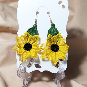 May include: A pair of sunflower earrings. Each earring features a yellow lace sunflower with a black center and green leaf. The earrings are displayed on a white card with a clear plastic stand.