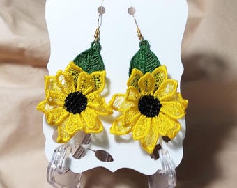 Puede incluir: Un par de pendientes de girasol. Cada pendiente presenta un girasol de encaje amarillo con un centro negro y una hoja verde. Los pendientes se exhiben en una tarjeta blanca con un soporte de plástico transparente.