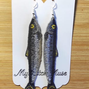 K&ouml;nnte beinhalten: Gestickte Fisch-Ohrringe mit silbernen Haken. Die Ohrringe zeigen ein detailliertes Design eines Fisches mit grauen und schwarzen Schuppen, gelben Flossen und gelben Augen. Die Ohrringe werden auf einer wei&szlig;en Karte mit dem Text "My Sketch Muse" pr&auml;sentiert.