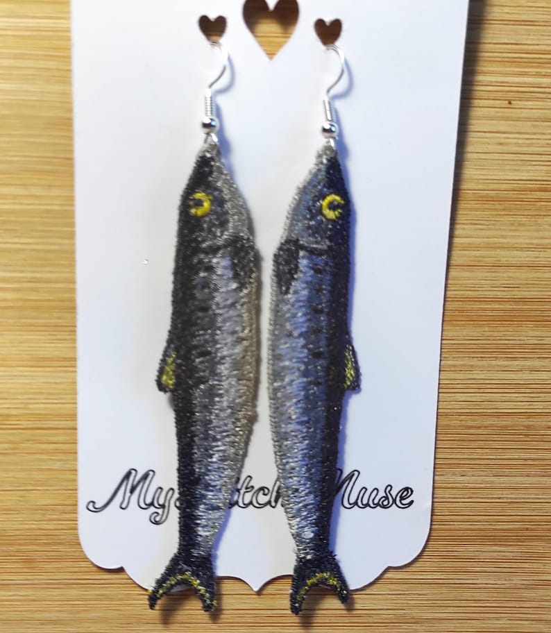 K&ouml;nnte beinhalten: Gestickte Fisch-Ohrringe mit silbernen Haken. Die Ohrringe zeigen ein detailliertes Design eines Fisches mit grauen und schwarzen Schuppen, gelben Flossen und gelben Augen. Die Ohrringe werden auf einer wei&szlig;en Karte mit dem Text "My Sketch Muse" pr&auml;sentiert.