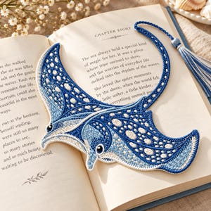 May include: A manta ray shaped bookmark featuring intricate blue and white embroidery with a spotted pattern. The bookmark includes a long, thin tail that extends into a blue and white tassel, resting on an open book page.
