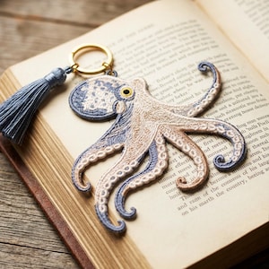 Puede incluir: Un llavero con un parche bordado de pulpo en tonos azul y beige con un ojo amarillo. El pulpo está unido a una anilla de metal dorado que también sostiene una borla de tela azul a juego con una tapa de color plateado.