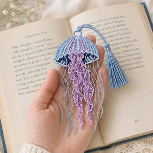 Peut inclure: Marque-page artisanal en forme de méduse, avec un design bleu et violet. Le marque-page est orné d'un pompon et est fabriqué à partir de fils tissés. Le motif de la méduse est complexe et détaillé, avec de longs tentacules fluides. Il est posé sur un livre ouvert.