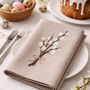 Puede incluir: Servilleta de lino beige con un bordado de ramas de sauce. La servilleta está doblada sobre un plato blanco. Una cesta de huevos de Pascua y un pastel están en el fondo, sugiriendo un tema festivo.