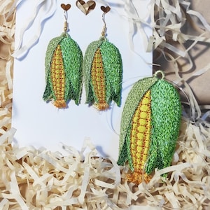 Puede incluir: Pendientes y colgante de maíz bordados. Los pendientes y el colgante presentan un diseño detallado de mazorcas de maíz, con hojas verdes y granos amarillos. Los pendientes cuelgan de ganchos dorados.