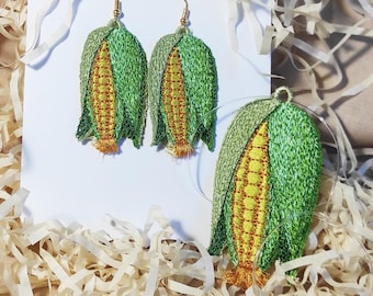 May include: Embroidered corn earrings and a matching pendant. The earrings and pendant feature a detailed design of corn on the cob, with green husks and yellow kernels. The earrings hang from gold-colored hooks.