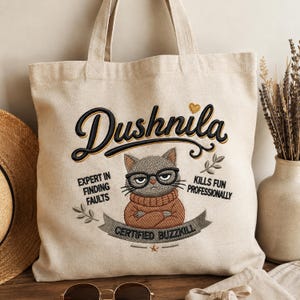 Puede incluir: Un bolso de tela beige con un diseño bordado de un gato gruñón con gafas y un suéter marrón. El texto dice Dushnila, Expert in finding faults, Kills fun professionally y Certified buzzkill, acompañado por un pequeño corazón bordado.