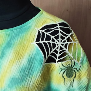 Puede incluir: Una camiseta teñida con tintes de amarillo y verde azulado, con un diseño de telaraña blanca en el hombro. Una araña verde está bordada debajo de la telaraña. La camiseta tiene un cuello negro.