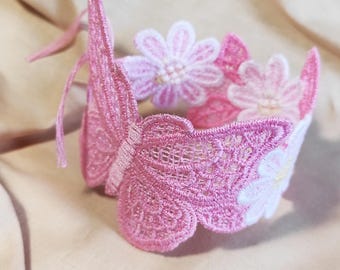 Puede incluir: Una corona rosa de encaje con mariposas y flores. La corona presenta una mariposa en un lado y varias flores bordadas. La corona está hecha de encaje rosa y tiene una cinta.