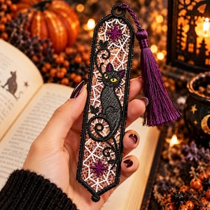 May include: A rectangular embroidered bookmark featuring a black cat with yellow eyes, surrounded by a white web-like pattern and purple star accents. The bookmark is framed in black thread and includes a long, deep purple silk tassel at the top.