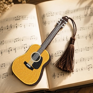 Könnte beinhalten: Ein gitarrenförmiges Lesezeichen mit gelber und brauner Stickerei und einer dunkelbraunen Quaste am Kopfstück. Das Lesezeichen ist mit einer strukturierten Garnoberfläche gefertigt und zeigt ein detailliertes Schallloch- und Griffbrettdesign.