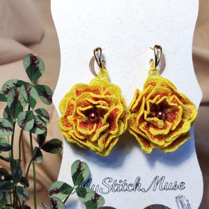 Può includere: Un paio di orecchini floreali fatti a mano. Ogni orecchino presenta un fiore ricamato giallo e rosso con un centro di perline marroni. Gli orecchini hanno ganci color oro. Lo sfondo include una carta bianca con il testo "My Stitch Muse".