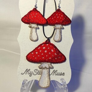 Op de afbeelding: Een rode sieradenset met paddenstoelthema, bestaande uit een ketting en bijpassende oorbellen. De paddenstoelkapjes zijn rood met witte stippen en de stelen zijn wit. De set wordt gepresenteerd op een witte kaart met de tekst "My Sti Muse".