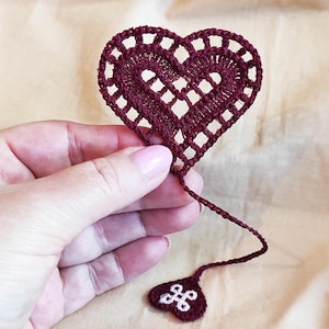 Può includere: Segnalibro a forma di cuore all'uncinetto bordeaux con un design traforato. Il segnalibro ha un lungo e sottile stelo che conduce a un piccolo tag a forma di cuore con un motivo decorativo bianco. L'articolo è tenuto su uno sfondo neutro.