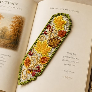 May include: An elongated, pointed bookmark featuring intricate white lace embroidery with a green border. The design includes yellow maple leaves, brown oak leaves, small red berries, and two acorns, all connected by swirling tan embroidered stems.