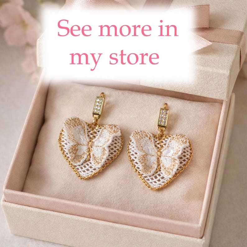 Puede incluir: Pendientes en forma de coraz&oacute;n con dise&ntilde;o de mariposa, bordados en blanco y dorado. Los pendientes se muestran en una caja de regalo con interior rosa y una cinta rosa. El texto "See more in my store" es visible.