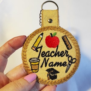 May include: A beige felt keychain with gold trim, featuring a red apple, pencil, ruler, scissors, coffee cup, and graduation cap. The text "Teacher Name" is embroidered in black. Includes a metal keyring.