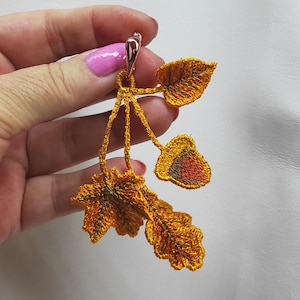 Könnte beinhalten: Ein goldfarbener Ohrring in Blattform mit aufwendiger Stickerei. Der Ohrring hat drei Blätter mit detaillierten Adern und einen Hakenverschluss. Die Blätter sind in Gold-, Orange- und Grüntönen gehalten.