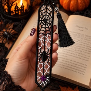May include: A black and white embroidered bookmark featuring a spider web pattern, a central black spider, and a small purple star at the bottom. The bookmark has a rounded top with a black tassel attached by a silver-toned bead and a black cord.