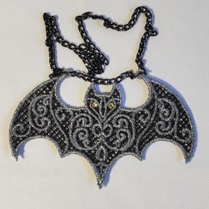 Può includere: Una collana a forma di pipistrello nera e argento. Il ciondolo presenta intricati dettagli simili a pizzo ed è impreziosito da due gemme iridescenti per gli occhi. La collana ha una catena nera.