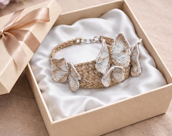 Puede incluir: Una pulsera dorada con detalles de mariposas bordadas, presentada en una caja de regalo beige con forro de satén. La pulsera presenta un diseño tejido y un cierre plateado. La caja está atada con una cinta a juego.