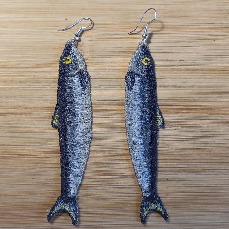 A pair of freestanding lace embroidered sardine earrings in shades of gray, black, and yellow.