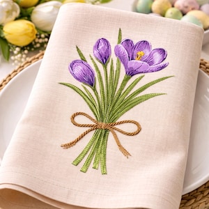Op de afbeelding: Een beige linnen servet geborduurd met een boeket paarse krokusbloemen en groene stelen, vastgebonden met een bruine touwstrik. Het servet is opgevouwen en op een wit bord geplaatst, met gele tulpen en paaseieren op de achtergrond.