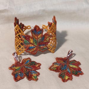 May include: A jewelry set featuring a cuff bracelet and two pendant earrings. The pieces are embroidered with a leaf design in shades of red, orange, yellow, green, and blue. The bracelet has a gold-colored openwork design.