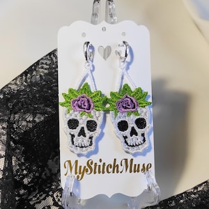 May include: Embroidered skull earrings with green leaves and purple flowers. The white skulls have black eyes and a smiling mouth. The earrings hang from silver hoops and are displayed on a white card with the text "MyStitchMuse".