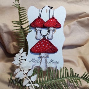 Op de afbeelding: Een rode sieradenset met paddenstoelthema, inclusief oorbellen en een hangerketting, gepresenteerd op een witte kaart. De paddenstoelen hebben rode doppen met witte stippen en witte stelen. De kaart heeft de tekst "My St* Muse".