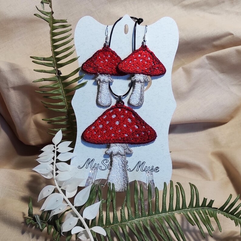Op de afbeelding: Een rode sieradenset met paddenstoelthema, inclusief oorbellen en een hangerketting, gepresenteerd op een witte kaart. De paddenstoelen hebben rode doppen met witte stippen en witte stelen. De kaart heeft de tekst "My St* Muse".