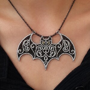 Può includere: Una collana con ciondolo a forma di pipistrello in pizzo nero con intricati ricami argentati e piccoli occhi gialli. Il ciondolo è sospeso a una sottile catena di metallo nero e presenta dettagliati motivi a volute sulle ali e sul corpo.