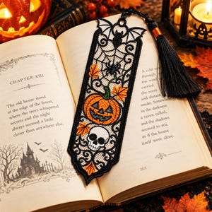 May include: A black lace bookmark featuring an embroidered orange jack-o'-lantern, a white skull, a spider web, and a bat at the top. The bookmark is accented with orange autumn leaves and includes a black tassel attached to the top with an orange band.