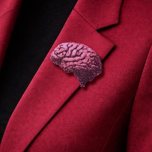 Könnte beinhalten: Ein weinrotes Sakko mit einer glitzernden, rosa geformten Gehirnbrosche, die an der Revers befestigt ist. Die Brosche verleiht der formellen Kleidung einen Hauch von Laune. Der Hintergrund ist ein schwarzes Hemd.